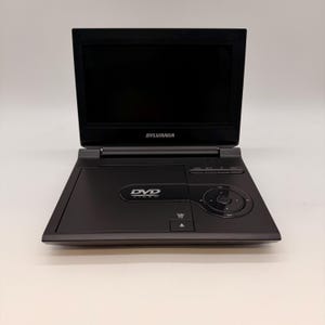 以下が含まれることがあります： 黒いSylvaniaポータブルDVDプレーヤー、スクリーン付き。DVDプレーヤーには、内蔵スクリーンとコントロールボタンがあります。DVDロゴが前面に表示されています。スクリーンは開いており、黒い画面が表示されています。