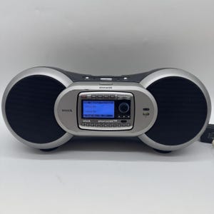 May include: A silver and black Sirius Sportster radio with two large black speaker grilles on either side. The radio has a blue screen displaying text and a circular control knob. The word "Subscribe" is visible on the screen.