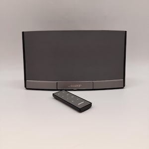 Puede incluir: Un altavoz Bose gris oscuro con un mando a distancia a juego. El altavoz tiene forma rectangular con una parte frontal de malla y un borde plateado. El mando es negro con botones y el logotipo de Bose. Aproximadamente 30 cm de ancho.