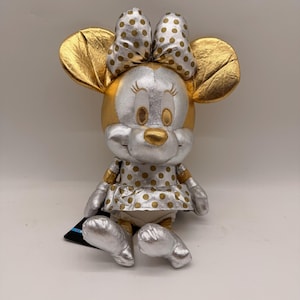 May include: A plush Minnie Mouse doll with gold and silver metallic accents. The doll features gold ears, a gold nose, and a polka-dot bow. The dress and shoes are silver with gold polka dots. A black tag is attached to the doll.
