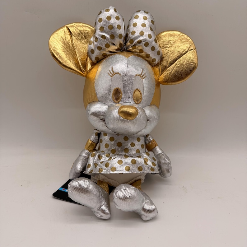 May include: A plush Minnie Mouse doll with gold and silver metallic accents. The doll features gold ears, a gold nose, and a polka-dot bow. The dress and shoes are silver with gold polka dots. A black tag is attached to the doll.