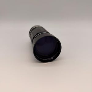 Puede incluir: Un objetivo de cámara negro con un interior morado oscuro. El objetivo tiene un agarre estriado y una banda plateada. El texto "Lens Made in Japan" es visible en el objetivo. El objetivo está sobre un fondo blanco.