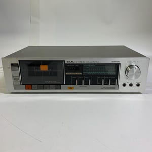 May include: A silver TEAC V-44C Stereo Cassette Deck. The deck has a cassette tape compartment, a display, and various buttons and knobs. The text "TEAC V-44C Stereo Cassette Deck" is printed on the front.