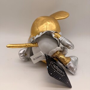 May include: A plush toy of a character with a gold head and ears, silver shoes, and a white body. The toy has a gold and silver outfit and a tag with the Disney logo.