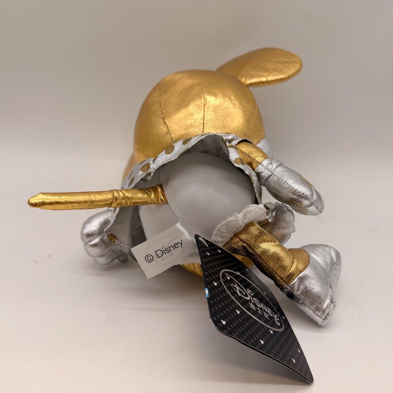 May include: A plush toy of a character with a gold head and ears, silver shoes, and a white body. The toy has a gold and silver outfit and a tag with the Disney logo.