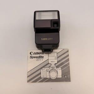 May include: A black Canon Speedlite 277T flash unit with a white and grey instruction manual. The flash has a rectangular head with a clear panel and a smaller dark grey panel. The manual features a line drawing of a camera.