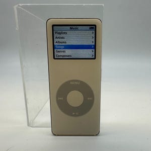 May include: A white digital music player with a rectangular screen displaying a menu with the text Music, Playlists, Artists, Albums, Songs, Genres, and Composers. Below the screen is a circular gray click wheel with navigation icons.
