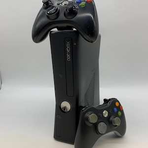 Peut inclure: Une console Xbox 360 noire avec deux manettes noires. La console a un bouton d'alimentation argenté et le texte "XBOX 360" à l'avant. Les manettes ont des boutons colorés et des pavés directionnels.