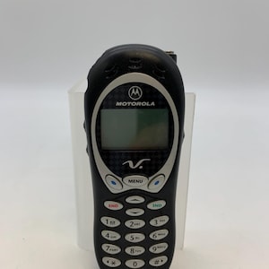 May include: Black Motorola 120c cell phone with a silver trim. The phone has a small screen, a keypad with numbers and function buttons, and the Motorola logo. The phone is in a vertical position.