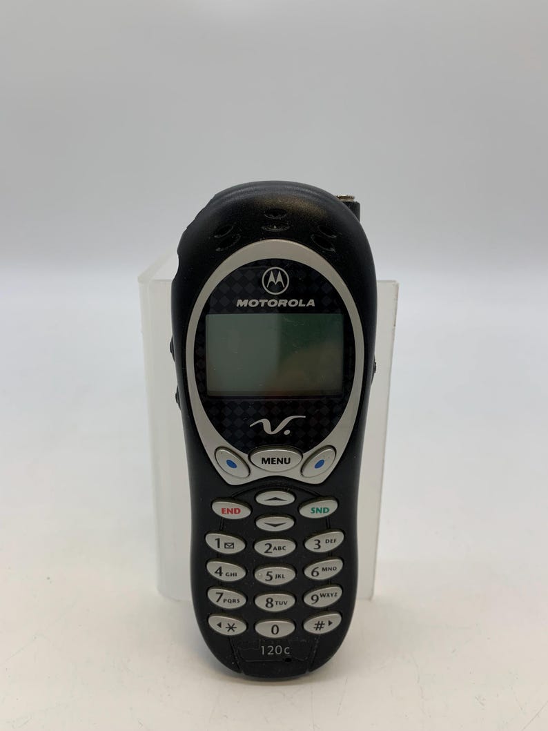 May include: Black Motorola 120c cell phone with a silver trim. The phone has a small screen, a keypad with numbers and function buttons, and the Motorola logo. The phone is in a vertical position.