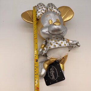 May include: A plush Minnie Mouse doll, primarily silver and gold, with a polka-dot dress and bow. The doll is approximately 15 inches tall. A Disney tag is attached. The doll's ears and bow are gold.