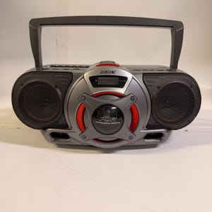 K&ouml;nnte beinhalten: Ein grauer, tragbarer Sony-Radiorekorder mit einem zentralen Power Drive Woofer mit roten Akzenten, einem digitalen Display und einem Tragegriff. Das Ger&auml;t verf&uuml;gt &uuml;ber zwei seitliche Lautsprecher mit schwarzen Gittern und dem Aufdruck PDW.