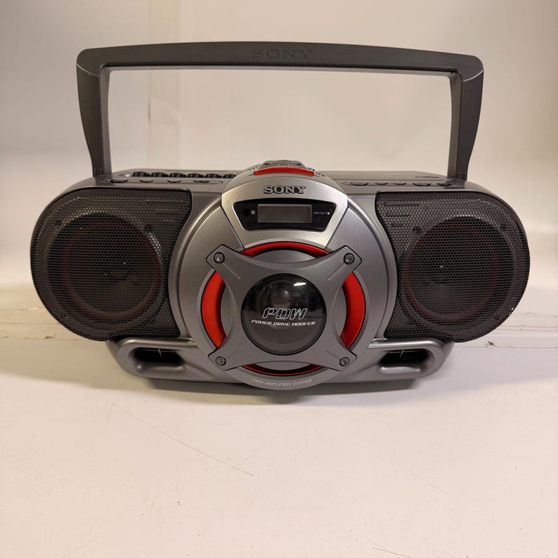 K&ouml;nnte beinhalten: Ein grauer, tragbarer Sony-Radiorekorder mit einem zentralen Power Drive Woofer mit roten Akzenten, einem digitalen Display und einem Tragegriff. Das Ger&auml;t verf&uuml;gt &uuml;ber zwei seitliche Lautsprecher mit schwarzen Gittern und dem Aufdruck PDW.