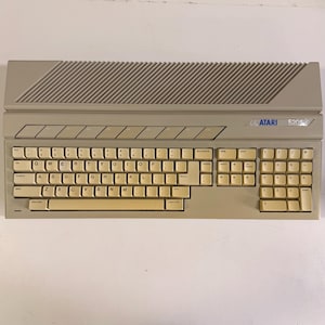 May include: A vintage Atari 520ST computer keyboard. The keyboard is beige with a full QWERTY layout, number pad, and function keys. The Atari logo and model number are visible on the top. The top section has a ridged design.