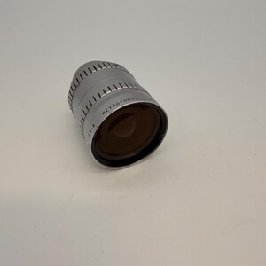 May include: A silver Retrofocus camera lens with a dark brown lens. The lens has a ridged design around the top and the text "RETROFOCUS" is printed on the side. The lens is angled slightly to the right.