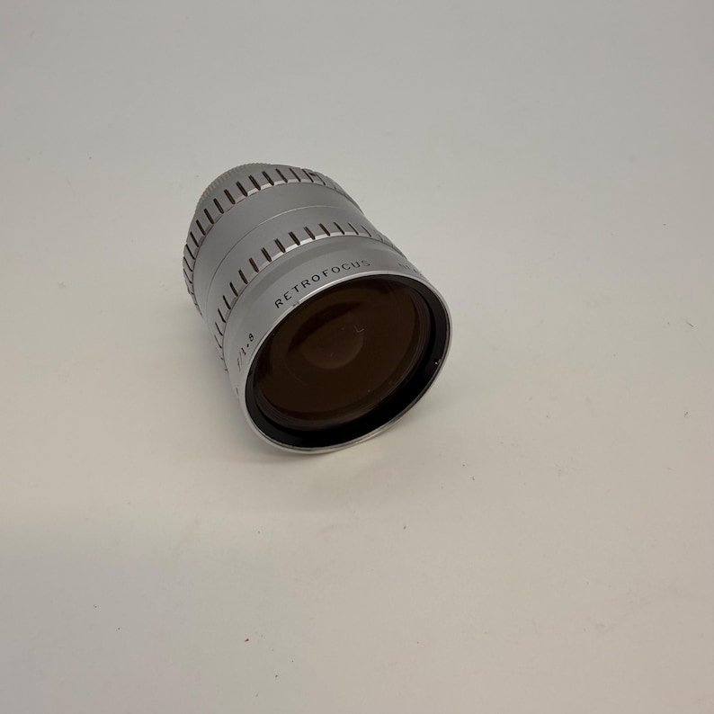 May include: A silver Retrofocus camera lens with a dark brown lens. The lens has a ridged design around the top and the text "RETROFOCUS" is printed on the side. The lens is angled slightly to the right.