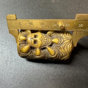 May include: A carved, golden-brown figurine of a reclining figure, approximately 1.5 inches long, is measured with a brass caliper. The figure has detailed facial features and ornate patterns. The background is a dark, textured surface.