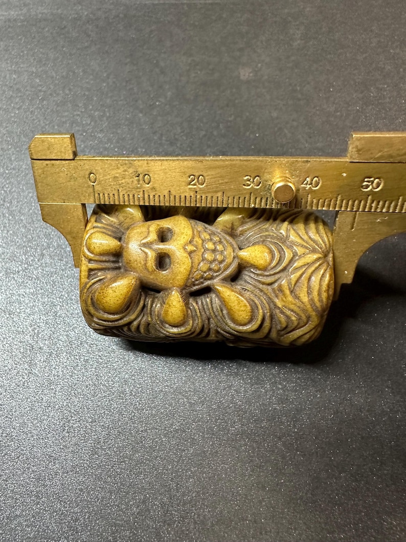 May include: A carved, golden-brown figurine of a reclining figure, approximately 1.5 inches long, is measured with a brass caliper. The figure has detailed facial features and ornate patterns. The background is a dark, textured surface.