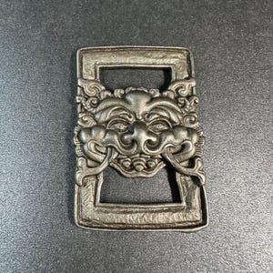 May include: A silver-colored rectangular belt buckle with a detailed, ornate design. The buckle features a central face with intricate facial features and decorative scrollwork. The buckle has two rectangular openings.