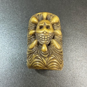 May include: A carved, rectangular sculpture with a smiling skull face. The sculpture is a light brown color and features detailed carvings of wavy lines and rounded shapes, giving it a textured appearance.