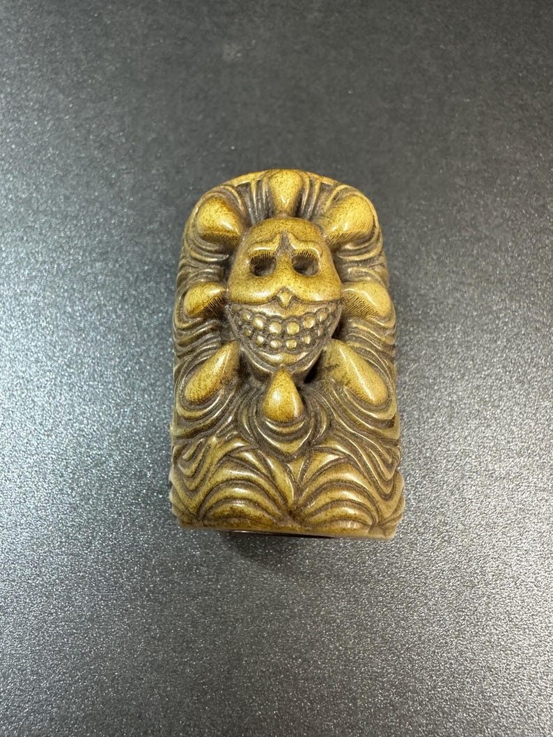 May include: A carved, rectangular sculpture with a smiling skull face. The sculpture is a light brown color and features detailed carvings of wavy lines and rounded shapes, giving it a textured appearance.