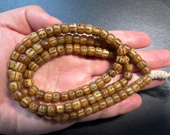 以下が含まれることがあります： 薄茶色のビーズの連なりで、濃い茶色のエッチングが施されています。ビーズは円筒形で、小さな点と線の繰り返し模様があります。連なりは、端が自然色の紐で結ばれています。