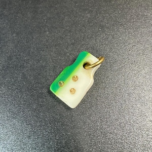 以下が含まれることがあります： 緑と白の配色が施された、小さな長方形のペンダント。上端は緑色で、白に変化します。3つの金色のスタッドが白い部分に埋め込まれています。上部には、吊り下げ用の金色のリングが付いています。