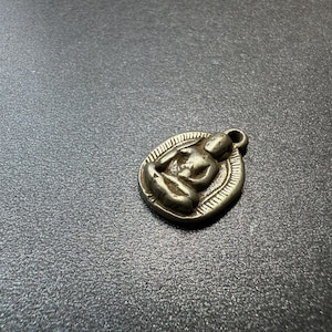 May include: A small, silver-colored pendant featuring a seated figure in a meditative pose. The pendant is oval-shaped with a textured border and a small loop at the top for a chain. The figure is detailed with visible facial features and clothing.