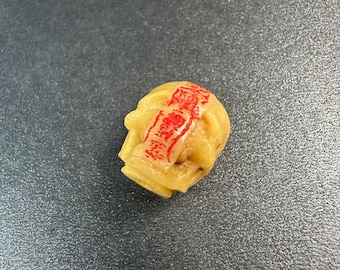 May include: A small, light-yellow carved object with red text on top. The object is in the shape of a head. The background is a dark gray surface.