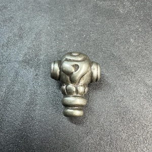May include: A metallic silver-toned bead with a rounded top featuring an embossed question mark symbol. The bead has a cylindrical base and two side protrusions, displaying a weathered, matte finish with visible dark grey recessed detailing.