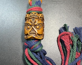 May include: A carved wooden pendant featuring a detailed face with multiple eyes and a knotted tassel. The tassel is made of red, green, and blue cords. The pendant has a loop at the top for hanging.