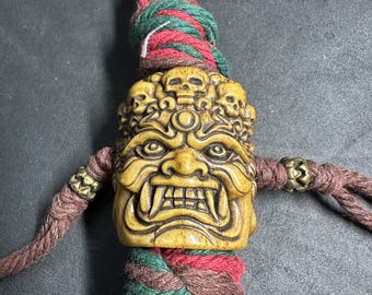 May include: A tan-colored carved bead featuring a fierce, fanged face with multiple skulls along the top edge. The bead is threaded onto a thick, braided cord in shades of red, green, and brown, accented with small, metallic, textured spacer beads.
