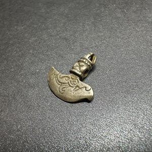 May include: A small, silver-colored pendant in the shape of a curved axe. The axe head features an engraved design, and the handle has decorative details. A small loop is at the top for attaching to a chain or cord. The pendant is likely made of metal.