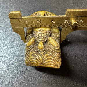May include: A carved, tan-colored figurine with a detailed face and textured body. A brass caliper is positioned over the figurine, measuring its width. The figurine appears to be a small sculpture or decorative item.