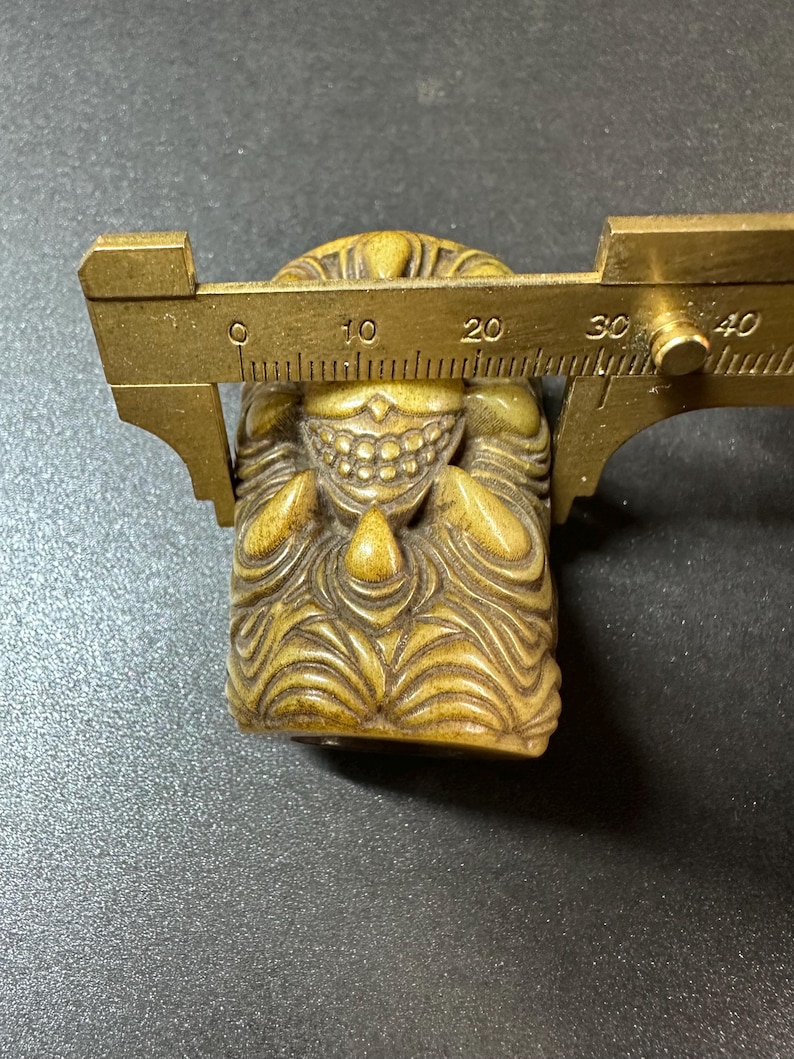 May include: A carved, tan-colored figurine with a detailed face and textured body. A brass caliper is positioned over the figurine, measuring its width. The figurine appears to be a small sculpture or decorative item.