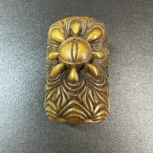May include: A rectangular, tan-colored carved object featuring a central eye motif surrounded by petal-like shapes. The lower portion of the piece displays a series of layered, wave-like patterns carved into the surface, creating a textured appearance.