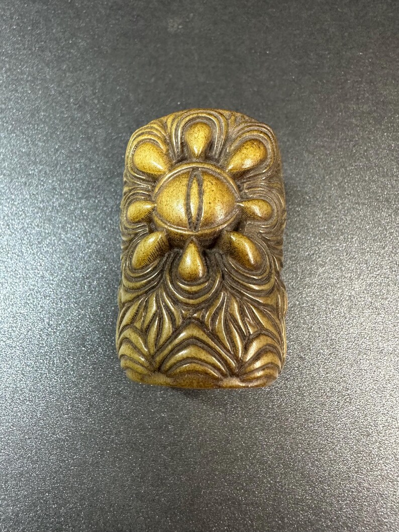 May include: A rectangular, tan-colored carved object featuring a central eye motif surrounded by petal-like shapes. The lower portion of the piece displays a series of layered, wave-like patterns carved into the surface, creating a textured appearance.