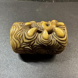 May include: A carved, cylindrical sculpture in shades of tan and brown. The sculpture features intricate details, including swirling patterns and a central design. The overall theme suggests a decorative or artistic object, possibly a Netsuke.