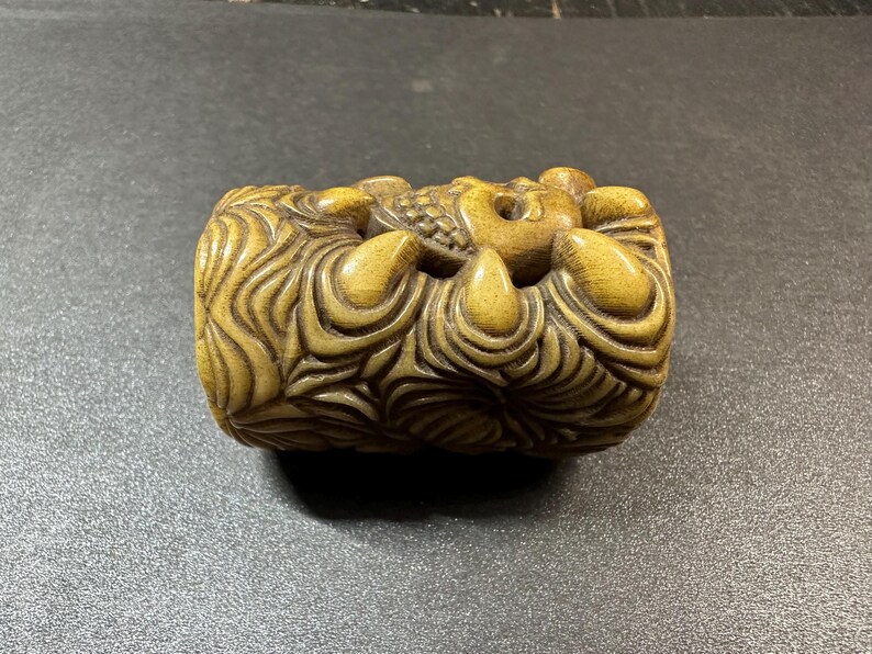 May include: A carved, cylindrical sculpture in shades of tan and brown. The sculpture features intricate details, including swirling patterns and a central design. The overall theme suggests a decorative or artistic object, possibly a Netsuke.