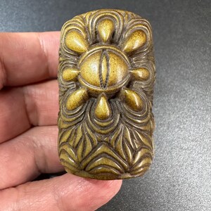 May include: A rectangular, brownish-gold carved object featuring a central eye motif surrounded by eight petal-like shapes. The surface is decorated with intricate, flowing, symmetrical relief patterns that extend across the entire piece.