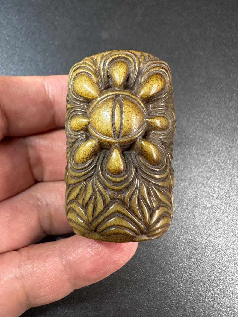 May include: A rectangular, brownish-gold carved object featuring a central eye motif surrounded by eight petal-like shapes. The surface is decorated with intricate, flowing, symmetrical relief patterns that extend across the entire piece.