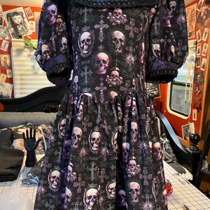 May include: A black dress with a purple floral collar and skull print. The dress features a drop waist, short sleeves, and a decorative trim. The skull pattern includes crosses and crowns.