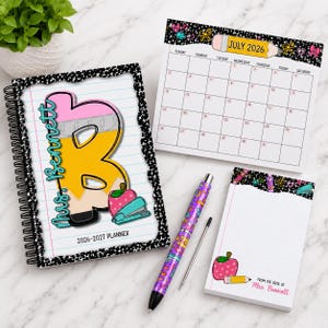 May include: A school-themed stationery set featuring a planner, calendar, notepad, and pen. The planner has a large letter "B" design with an apple and stapler. The calendar displays July 2026. The pen and notepad have matching designs.