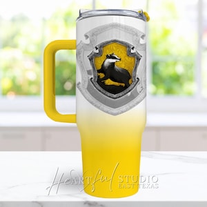 Puede incluir: Un vaso térmico amarillo y blanco con un escudo de tejón en blanco y negro. El vaso tiene un asa amarilla y una tapa transparente. La parte inferior del vaso es amarilla, desvaneciéndose a blanco en la parte superior.