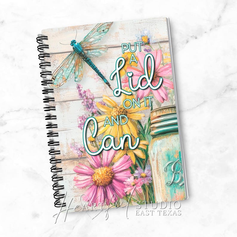 K&ouml;nnte beinhalten: Spiralgebundenes Notizbuch mit floralem Design und dem Text "Put a Lid on it and Can". Das Cover zeigt eine Libelle, G&auml;nsebl&uuml;mchen und ein Einmachglas auf einem verwitterten Holzgrund. Das Notizbuch stammt von Heartfelt Studio East Texas.