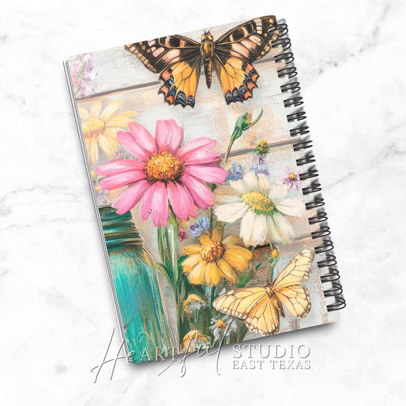 K&ouml;nnte beinhalten: Spiralgebundenes Notizbuch mit Blumen- und Schmetterlingsdesign. Das Cover zeigt eine rosa G&auml;nsebl&uuml;mchen, gelbe und wei&szlig;e Blumen und zwei Schmetterlinge vor einem rustikalen Holzhintergrund. Das Notizbuch hat eine t&uuml;rkisfarbene Einmachglas-Illustration und den Text "Heartful Studio East Texas".