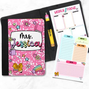 May include: A pink notebook with the name "Mrs. Jessica" in colorful letters, surrounded by stars, flowers, and book illustrations. A weekly focus planner and pen are also included.