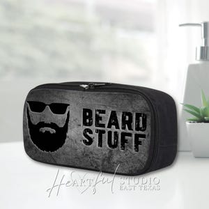 May include: Black rectangular travel bag with a zipper closure. The front features a graphic of a beard with sunglasses and the words "BEARD STUFF" in bold letters. The bag is designed for grooming products.