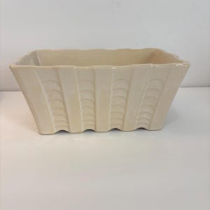 Puede incluir: Una maceta rectangular de cerámica color crema con un patrón vertical repetitivo de arcos semicirculares apilados en sus paredes exteriores. El recipiente tiene una abertura ancha y una base festoneada, con un acabado liso y brillante.
