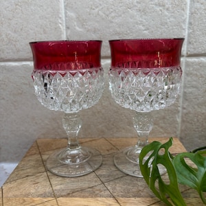May include: Two matching stemmed glasses featuring a deep red upper bowl and a clear, diamond-cut textured lower bowl. Each glass has a clear, faceted stem and a circular base, showcasing a distinct contrast between the colored rim and clear glass.